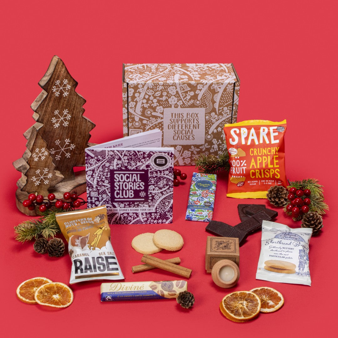 Festive Winter Gift Box open, showcasing sustainable treats including shortbread biscuits, chocolates, lavender candle, apple crisps, and nuts, with festive decorations like pine cones and dried oranges on a red background.