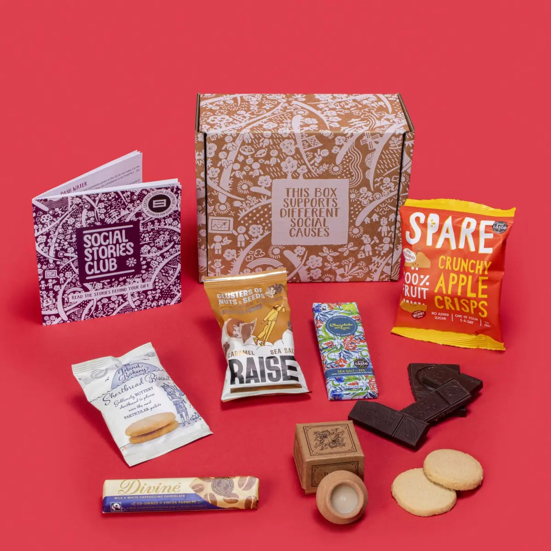 Festive Winter Gift Box open, showcasing sustainable treats including shortbread biscuits, chocolates, lavender candle, apple crisps, and nuts, with festive decorations like pine cones and dried oranges on a red background.