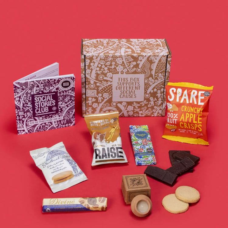 Festive Winter Gift Box open, showcasing sustainable treats including shortbread biscuits, chocolates, lavender candle, apple crisps, and nuts, with festive decorations like pine cones and dried oranges on a red background.