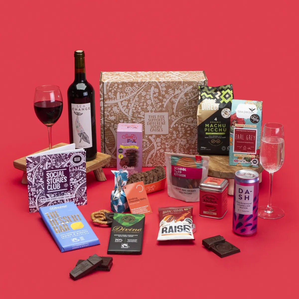 Indulgent Gift Box open, showcasing Earl Grey tea, Machu Picchu coffee, double chocolate cookies, white chocolate lemon cheesecake bar, fruit and nut dark chocolate, hot chocolate, chilli pretzels, maple pecan nuts, blackcurrant sparkling water, hot chocolate melts, wine, story booklet, and personalised note in eco-luxe packaging.