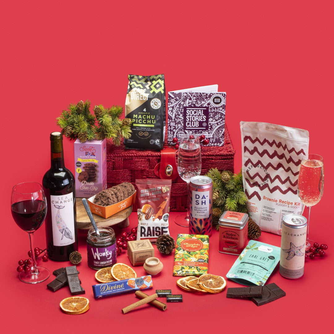 Indulgent Gift Hamper open, brimming with brownie kit, sparkling wine can, vegan cookies, Earl Grey tea, lavender candle, orange dark chocolate, salted caramel milk chocolate, raspberry sparkling water, napolitain, maple pecan nuts, onion relish, coffee beans, vegan hot chocolate, and story booklet in opulent sustainable packaging with subtle festive glow.