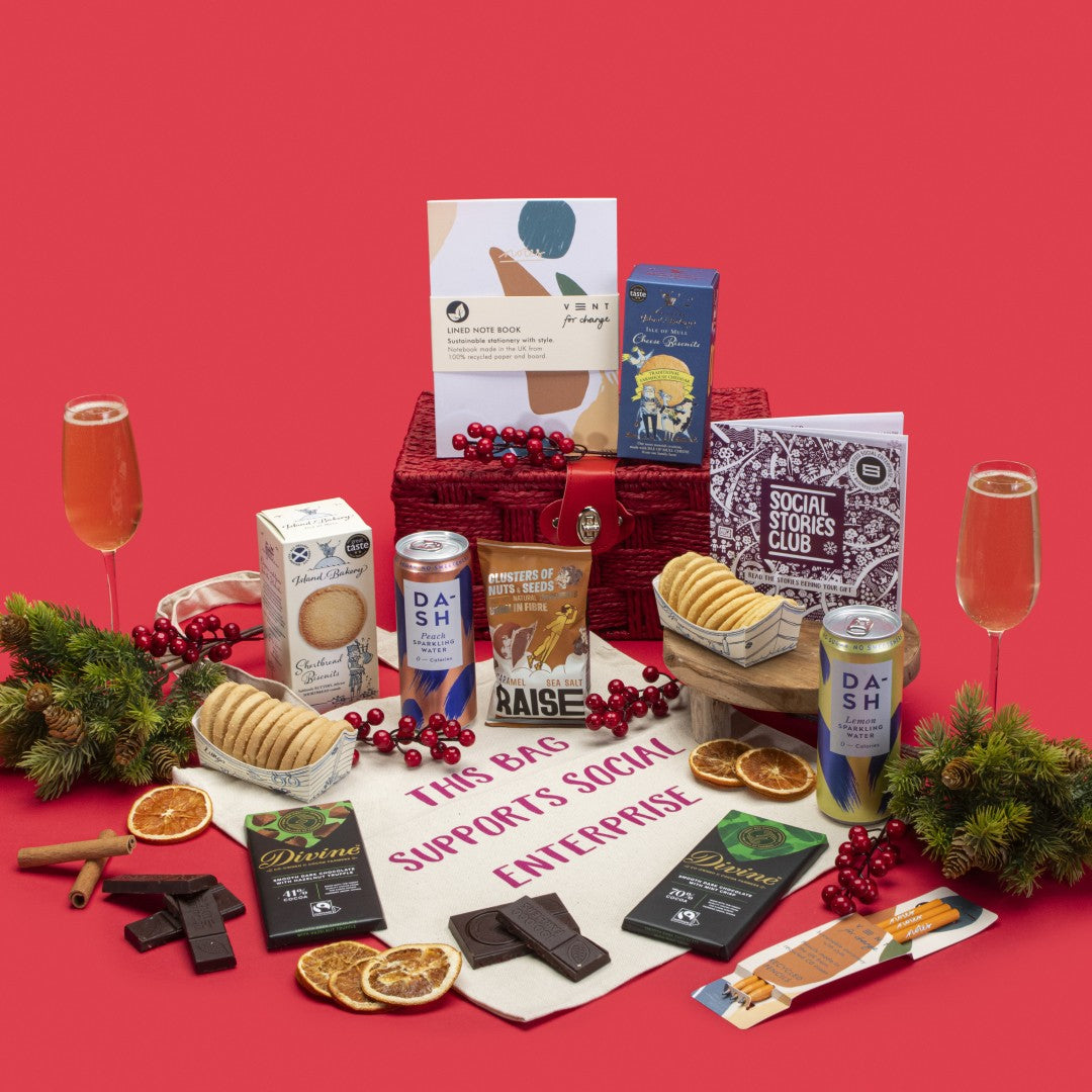 Let's Make Impact Gift Hamper open, showcasing shortbread and cheese biscuits, fruit and nut and mint crisp dark chocolates, salted caramel seeds and nuts, lemon and peach sparkling waters, cotton tote bag, A5 notebook, recycled pencil set, story booklet, and personalised note in empowering sustainable packaging with motivational festive accents.