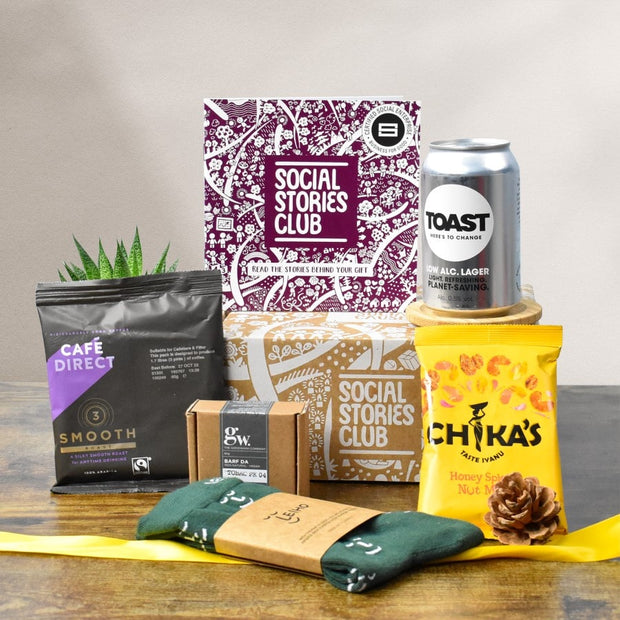 Sustainable Gift for Him. This is a sustainable gift box surrounded by products chosen for men such as a beard bar, beer, socks, and snacks. A story booklet is in this image which shares the social stories behind the products.